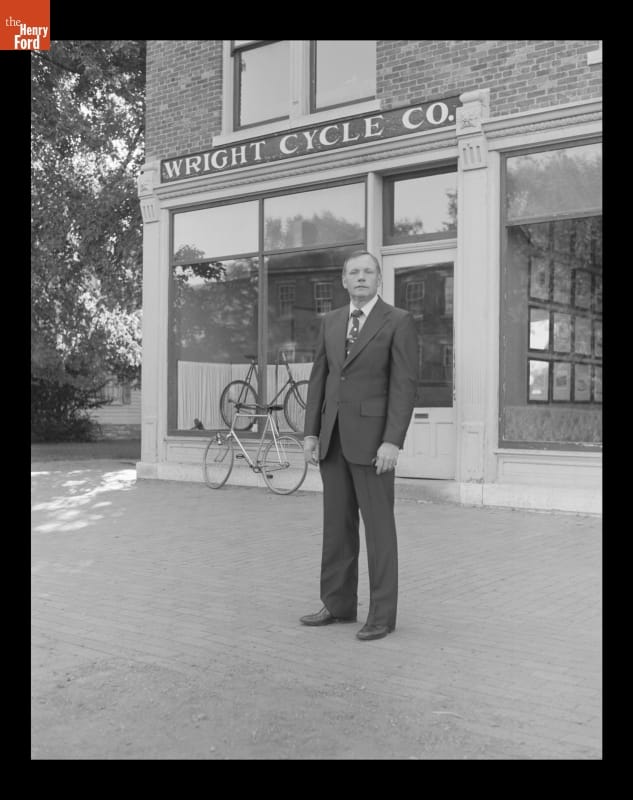 Neil Armstrong at the Wright Cycle Shop in Greenfield Village, August 16, 1979 Man in a suit stands before a brick building with a bicycle outside and one in the window