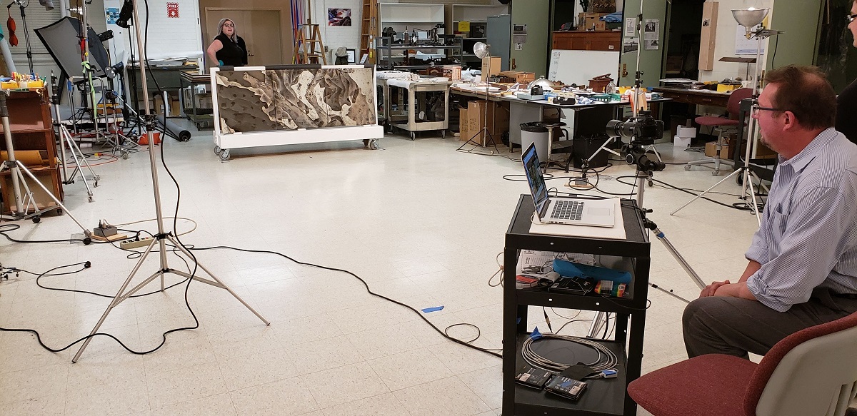 Photo Volunteer Doug Scoda assisting with photographing a mirrored panel in the former Photo Studio Man sits at cart with keyboard and woman stands with large mirror in cluttered room