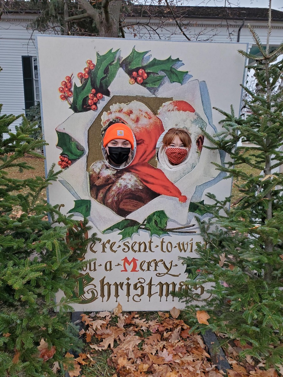 People pose behind a life-size postcard at Holiday Nights in Greenfield Village Two people stick their faces through holes in a life-size holiday postcard among evergreens