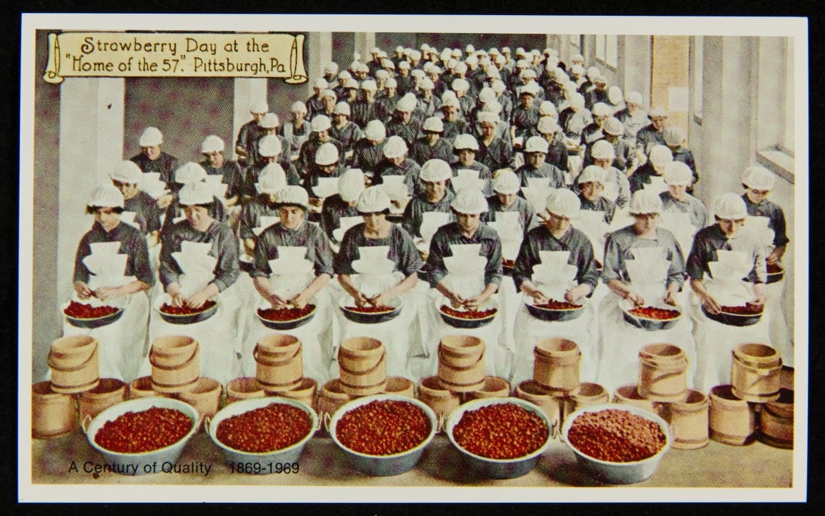 Postcard of a large group of women in white aprons and caps sorting strawberries, with large bowls of strawberries in the foreground