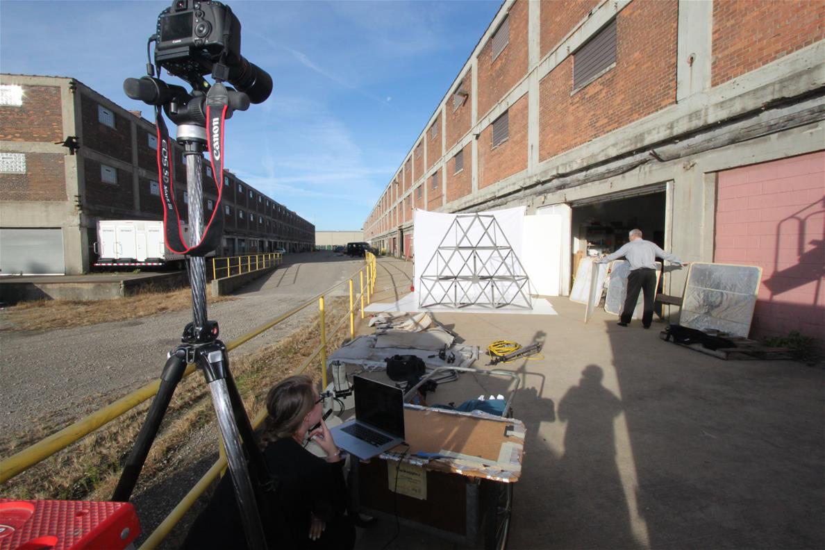 Digitizing a Large Artifact On-Site Camera on tripod in foreground pointed toward a large metal truss in front of white background, with large, low brick buildings extending into the distance on both sides
