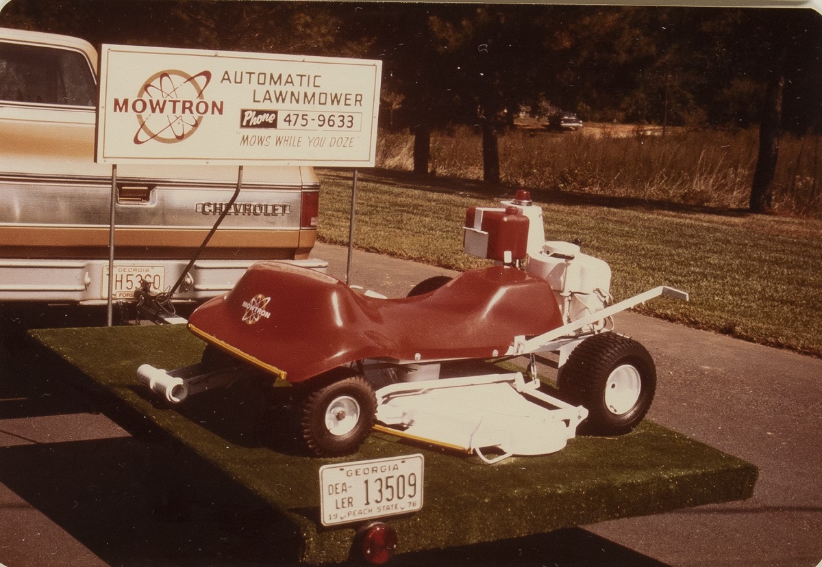 Binder of Documents Related to Mowtron Lawn Mower, 1974 / page 1 Lawmower on trailer behind truck, with sign containing text