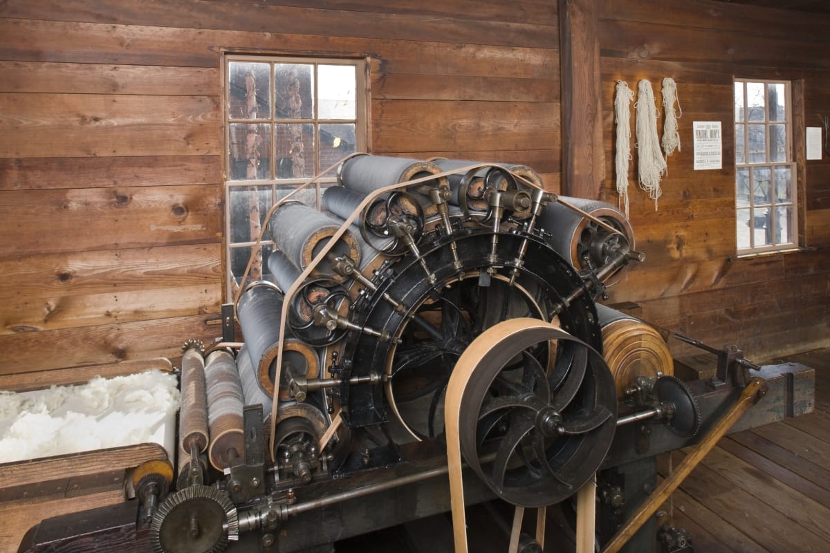 Carding Machine, 1850-1880 Machine with multiple rollers arranged in an arch shape with a conveyor belt filled with wool leading up to it