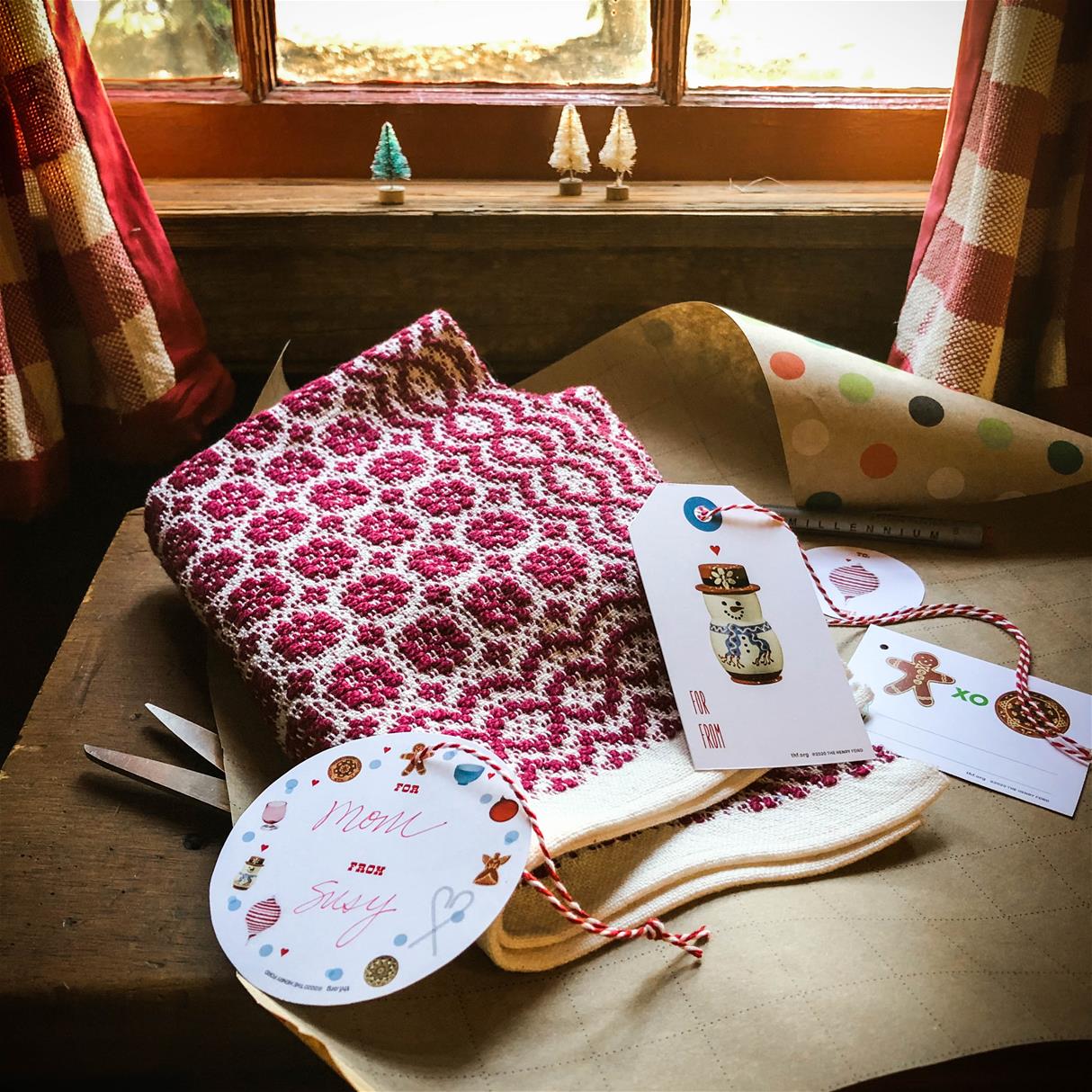Overshot Placemat, combined with THF-designed gift tags Red-and-white woven placemat on a piece of gift wrap with gift tags; window with red-and-white checked curtains and tiny Christmas trees on windowsill in background