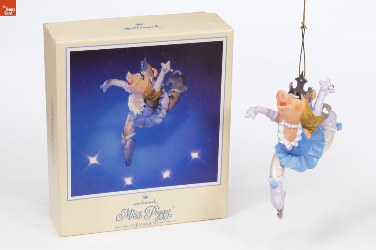 Hallmark "Miss Piggy" Christmas Ornament, 1983 Ornament of Miss Piggy in blue-and-white ice skating costume with long white gloves, arms and legs in dramatic pose, sitting next to box with image of the same ornament on the front