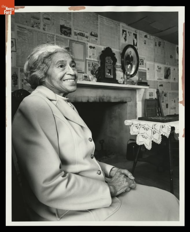 Woman with hair pulled back wearing suit jacket sits in chair with mantel and wall covered in newspaper behind her