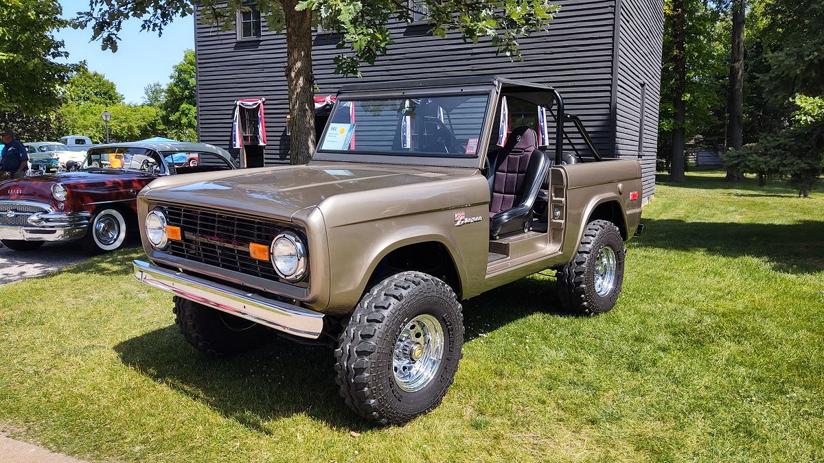 1977 Ford Bronco at Motor Muster 2022 in Greenfield Village Copper-colored Jeep-like vehicle parked among other vehicles in front of a gray wooden building