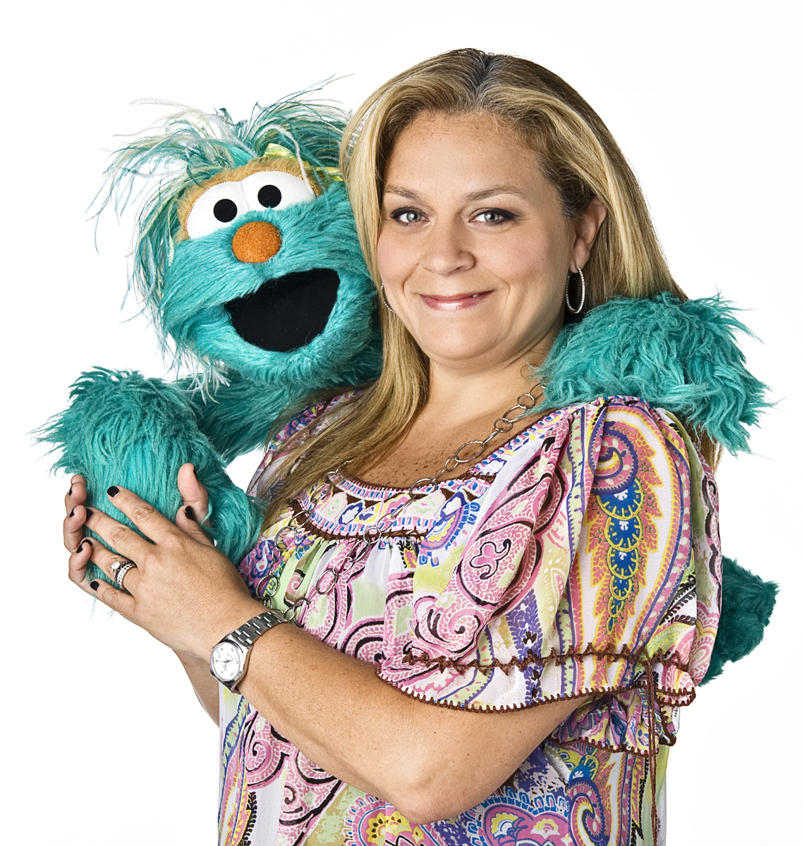 Carmen Osbahr-Vertiz and Rosita Smiling woman with long blonde hair stands with blue furry puppet over one shoulder