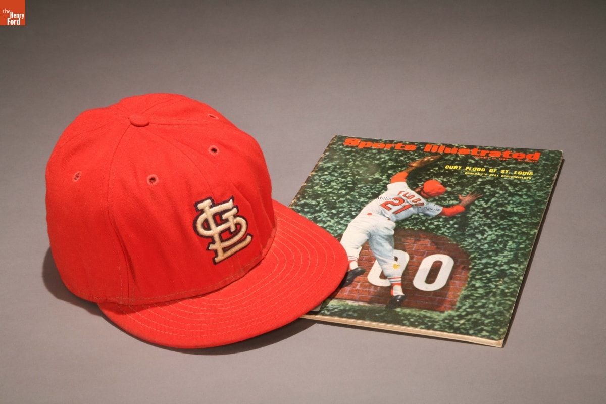 St. Louis Cardinals Baseball Cap Worn by Curt Flood, 1965-1969, and Sports Illustrated issue Red baseball cap with logo on front and Sports Illustrated magazine with image of baseball player jumping for ball on cover