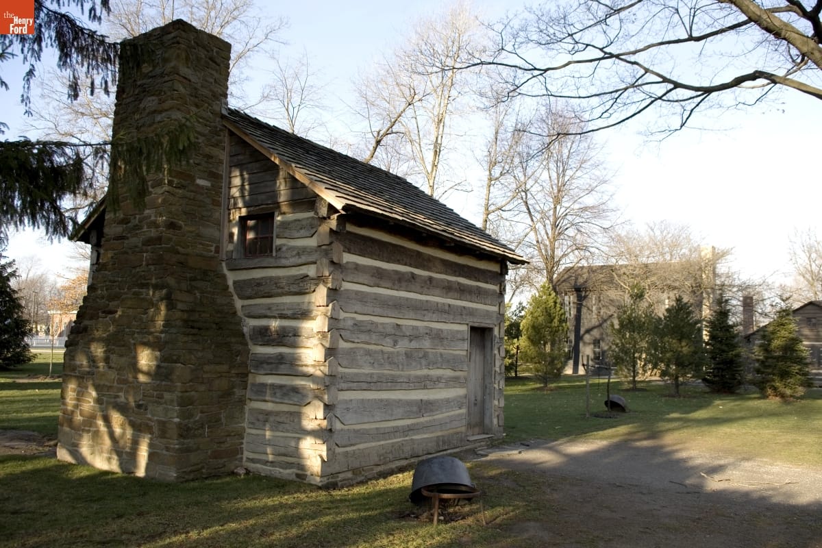 Log cabin with stone fireplace among trees with another building visible in the background