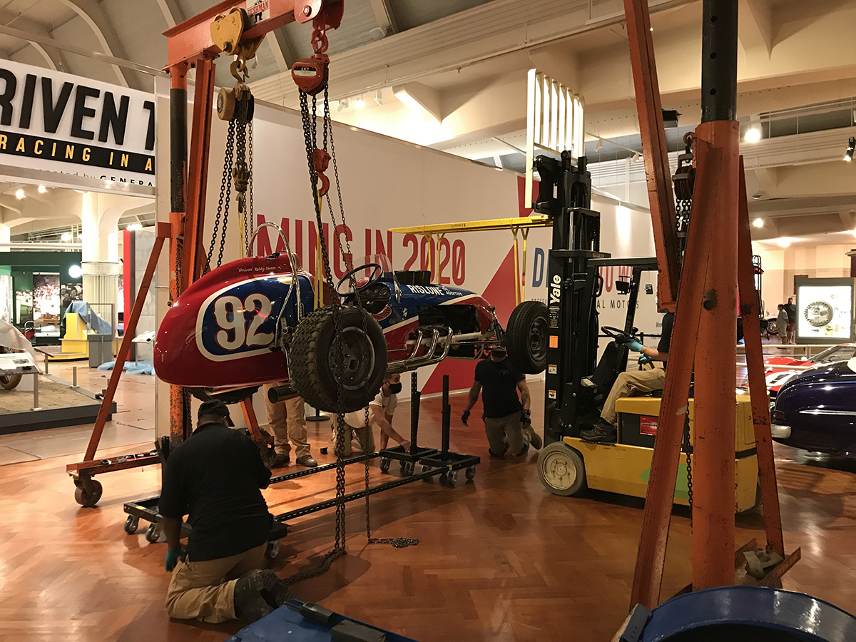 1958 Moore/Unser Pikes Peak Hill Climb racing car being lifted using a gantry and forklift. Red and blue race car suspended from a gantry and forklift with men kneeling by it