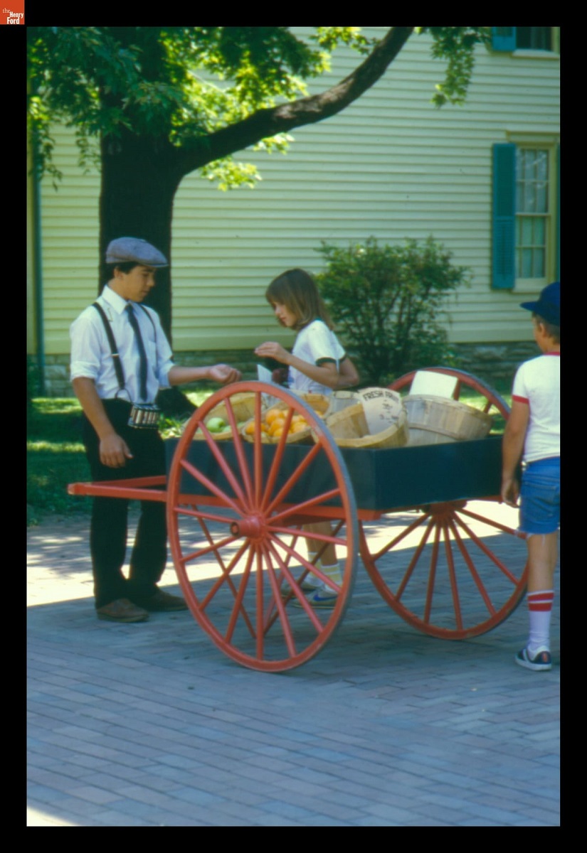 Street Vendor in Greenfield Village Man in suspenders and cap stands in front of cart filled with baskets, with two children nearby