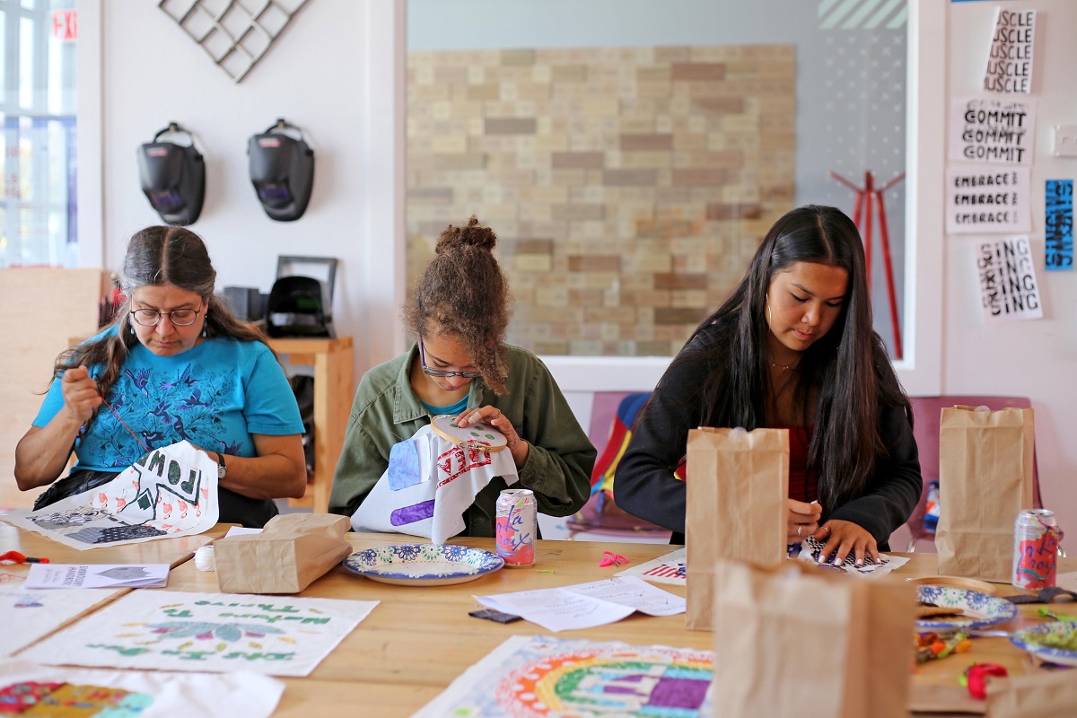 An SJSA embroidery volunteer sews along high school students during an embroidery workshop at Girls Garage Three people sew at a table full of drawings, plates, soda cans, and paper bags in a large, airy workspace