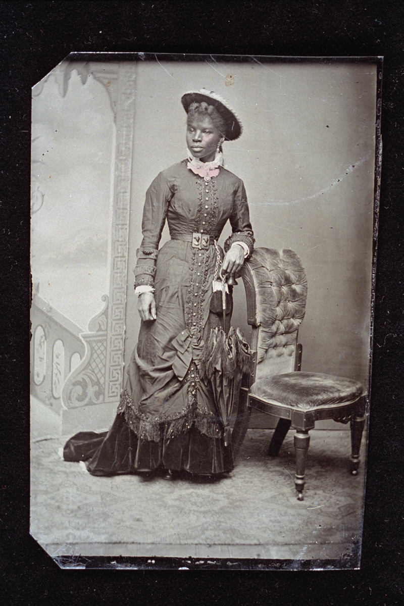 Woman Holding a Parasol, circa 1878 Black woman standing by a chair in front of a backdrop, wearing an elaborate dress and holding a parasol