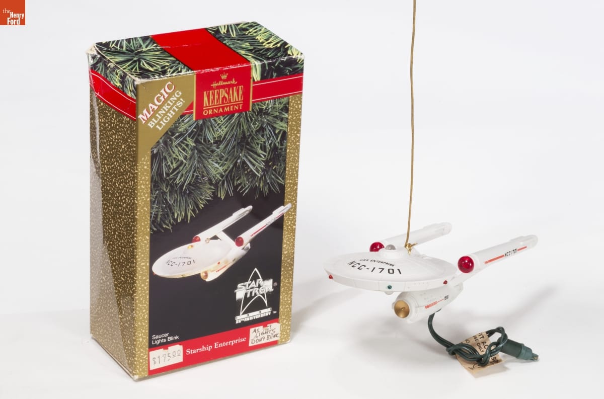 Hallmark "Starship Enterprise" Christmas Ornament, 1991 Ornament of spaceship with Christmas light extending from it, sitting next to gold-and-green box with image of the same ornament on the front