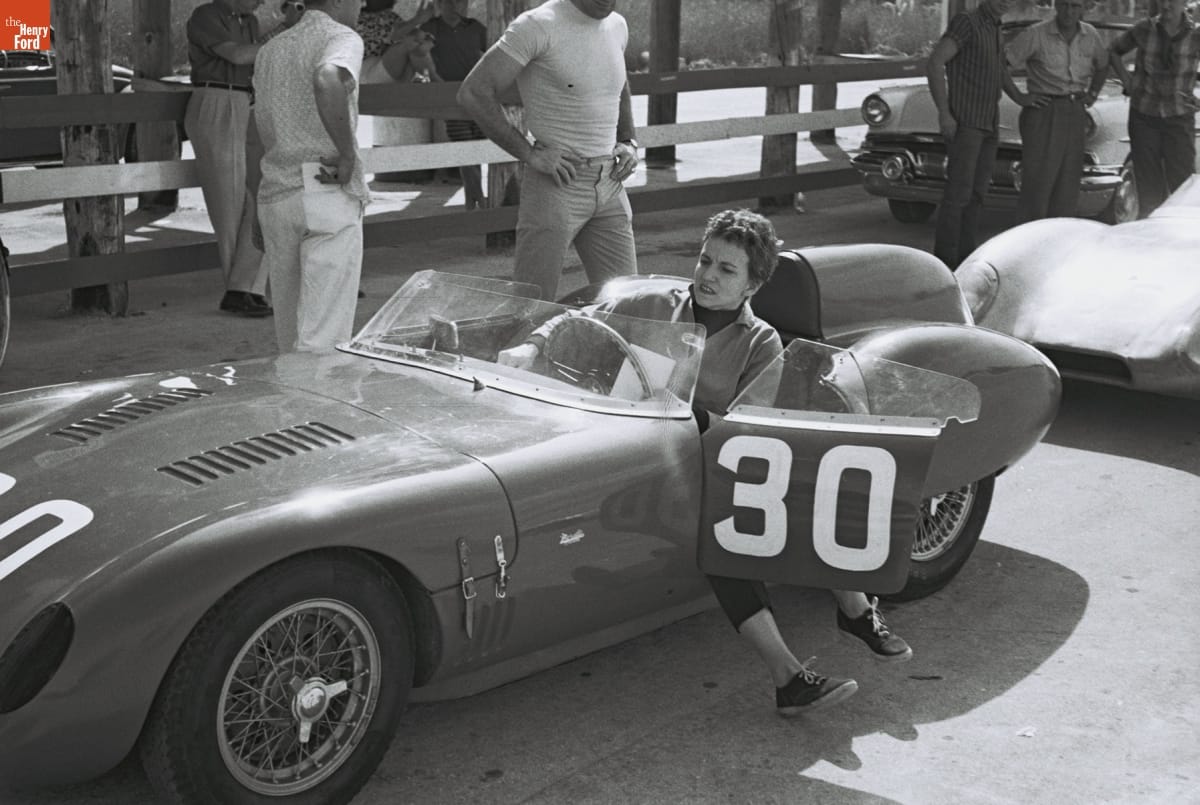 Denise McCluggage at Bahamas Speed Weeks, November-December 1959 Woman sits with legs out door of small, open race car among other cars and people