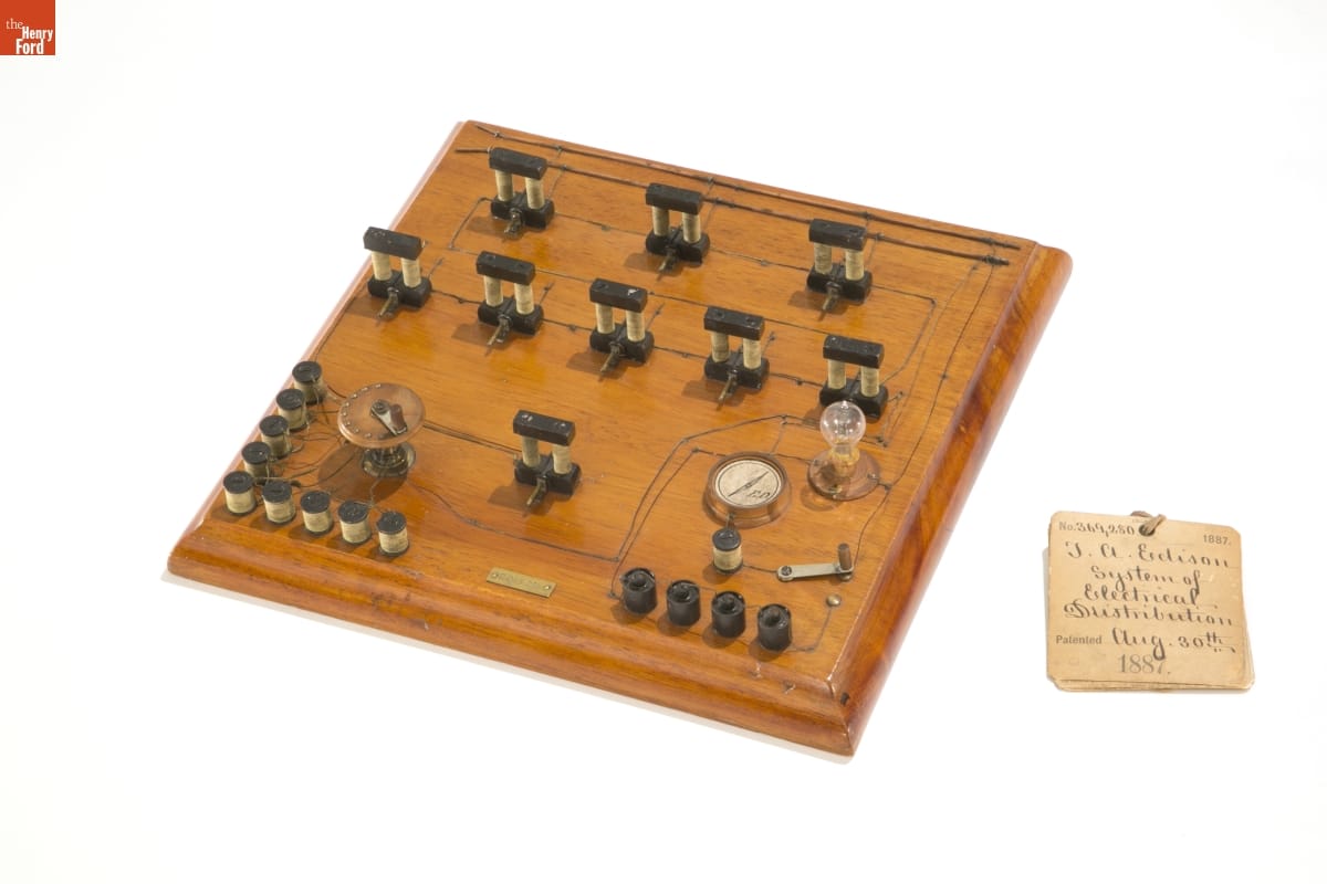 Edison Patent Model of Electric Distribution System, 1887 Wood board with small parts and wires attached to it; tag with handwritten text sits next to the board