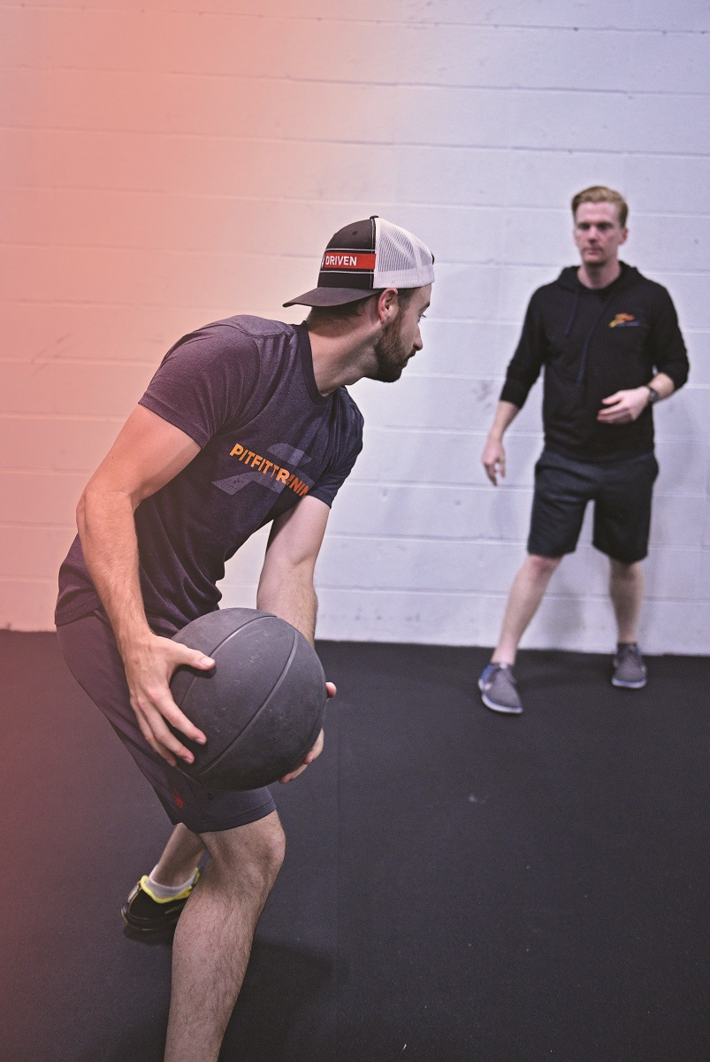 Driver James Hinchcliffe training at PitFit Man in shorts and t-shirt prepares to throw a medicine ball to another man in shorts and a hoodie