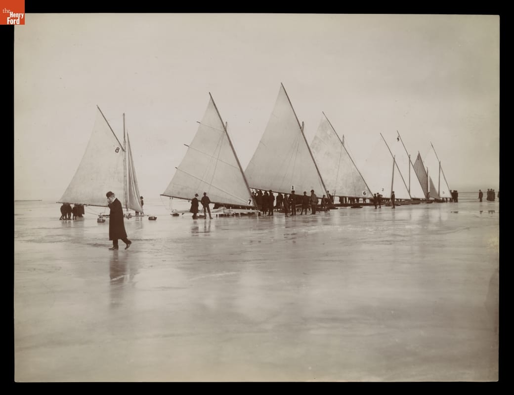 Conveyances with sails on ice with people standing around them