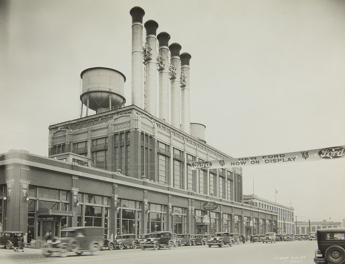 Exterior View, Ford Highland Park Plant, 30 March 1932 Factory topped with several smokestacks and a water tower, with cars driving by in front