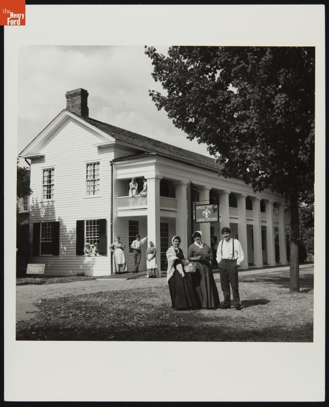 Historical Presenters outside Eagle Tavern in Greenfield Village, 1982 Black-and-white photo of people standing outside and on the first- and second-story balconies of a long wooden building with many columns