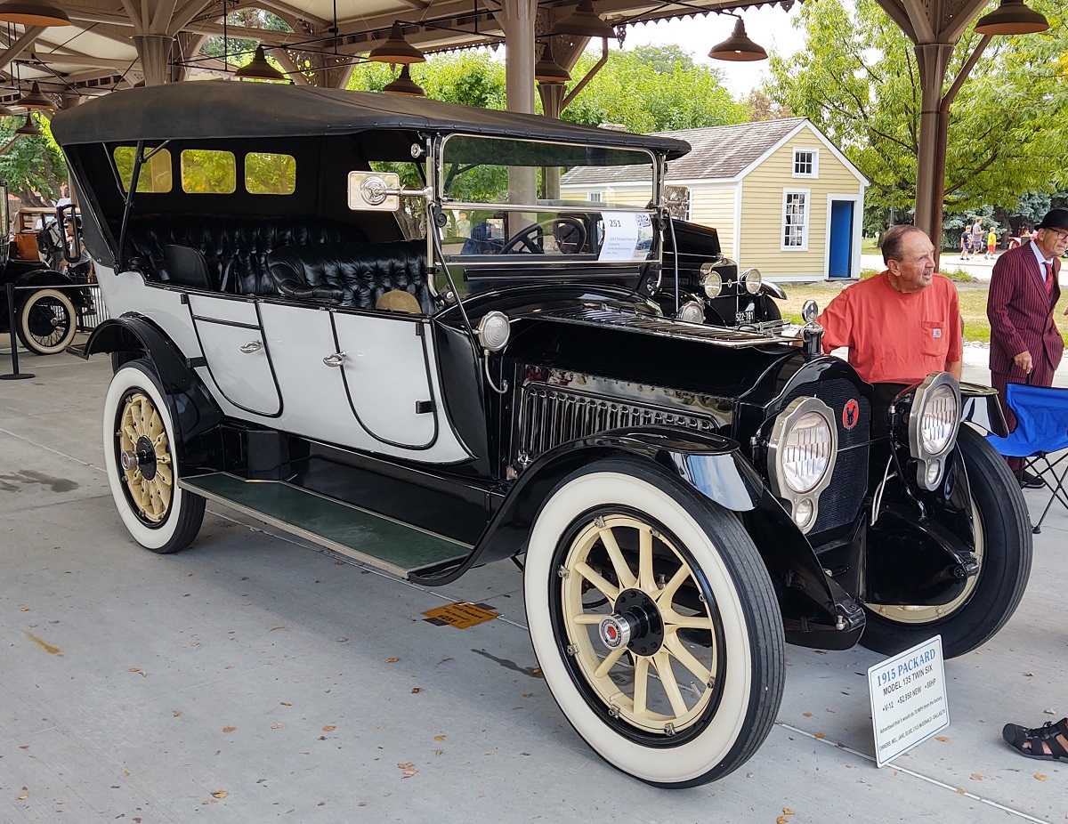 Old-fashioned car with white doors and black top, with open space where windows and dash would be, displayed in an open building with people nearby