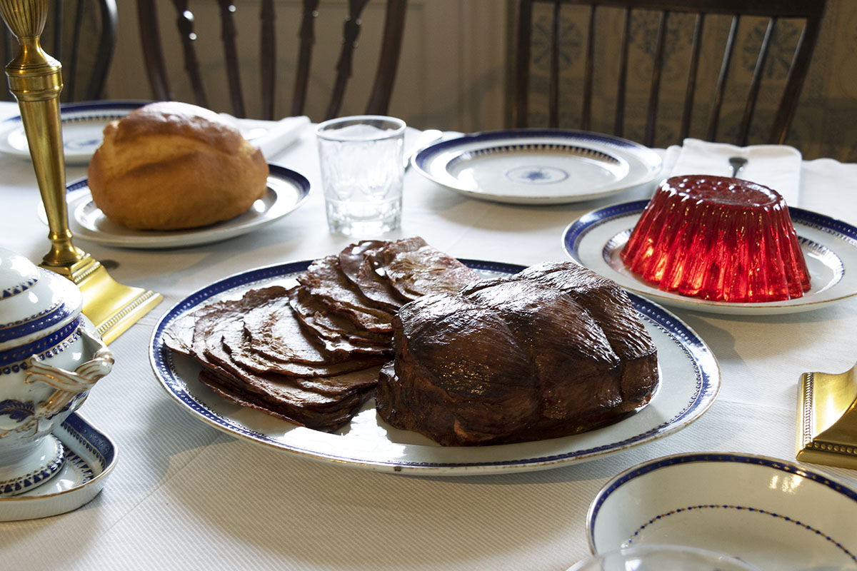 Meat roast (partially sliced), jello mold, and round loaf of bread on plates on table, with place settings nearby