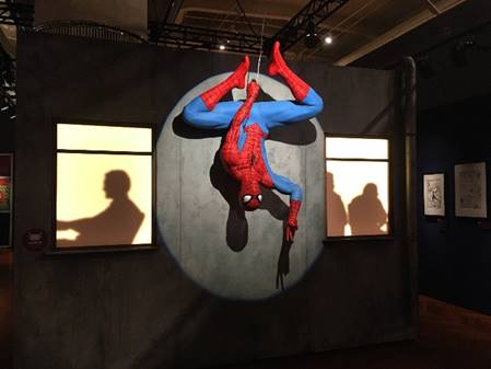 Spider-Man photo op in "Marvel: Universe of Super Heroes" exhibition Mannequin in blue and red unitard hanging upside down in spotlight with silhouettes of people in windows on either side