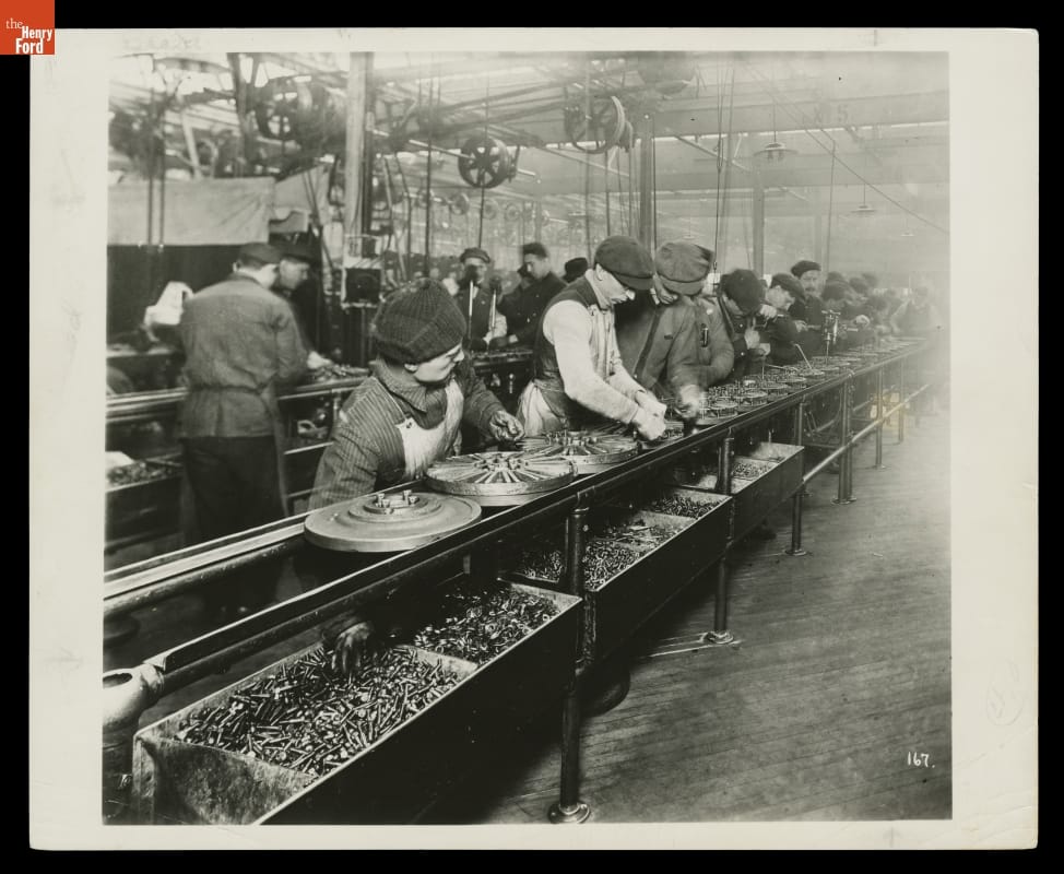 Magneto Assembly at the Ford Highland Park Plant, 1913 People work in a factory at a long line containing round parts, with bins of screws or bolts underneath