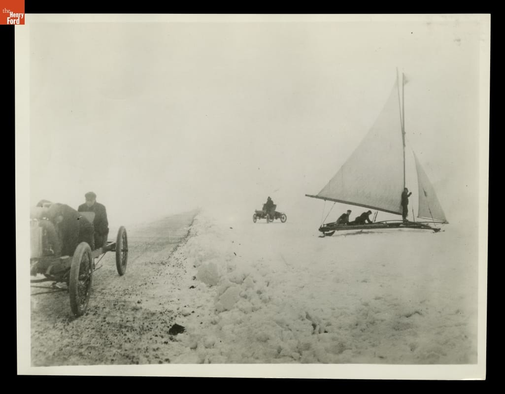 Car with two people in it on road or path on left side; on right side a conveyance with a sail and another car, each with people in them, sit in snow