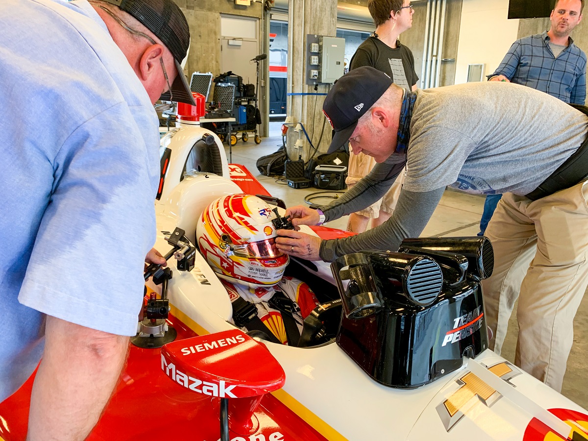 Setting up to film Joseph Newgarden at the Indy 500. One person adjusts a camera on the helmet of another person sitting in an open cockpit race car; additional people stand nearby