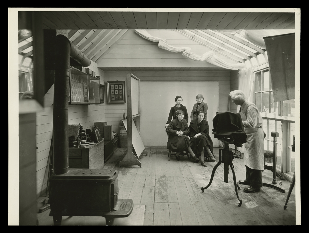 Photographer Charles Tremear Preparing for a Group Portrait in the Greenfield Village Tintype Studio, March 1935 Room with photographer at camera pointed at four women posing; woodstove in foreground and images on walls