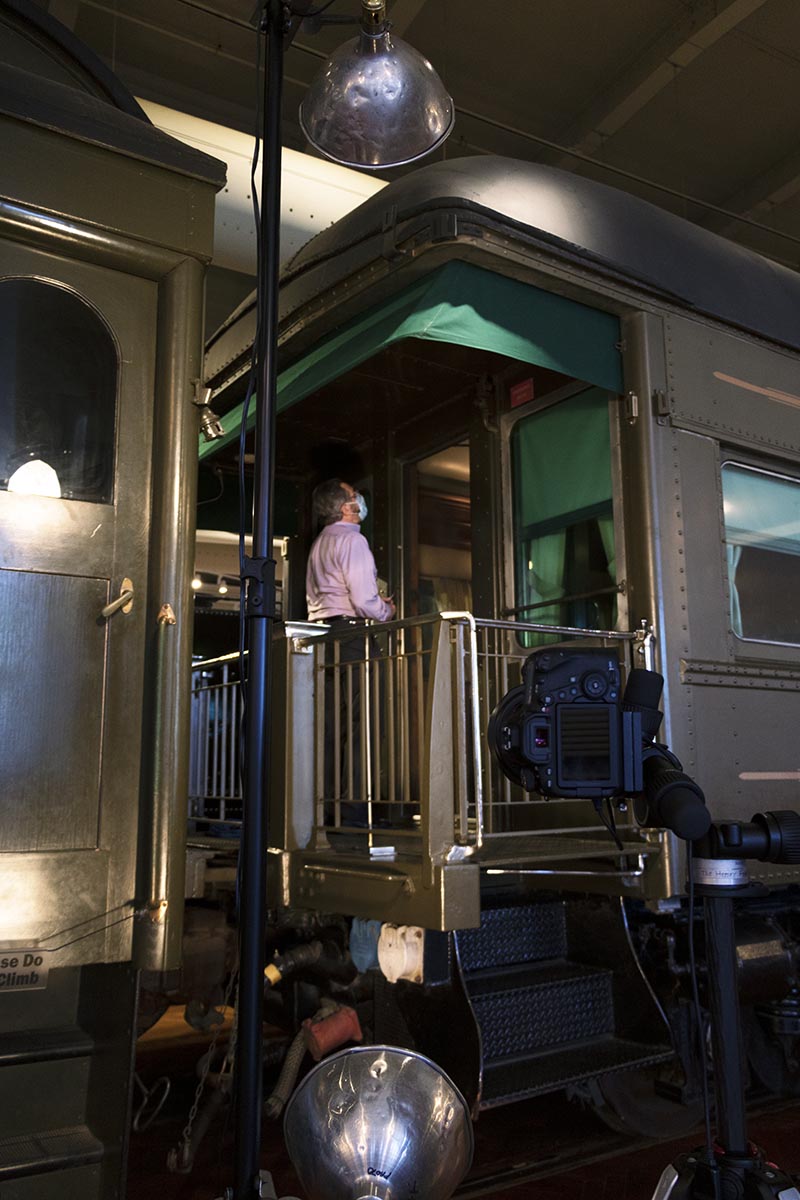 Photographer Rudy Ruzicska setting up lights for our first exterior shot of the railcar Man standing on back platform of railcar in large room, with camera and light in foreground