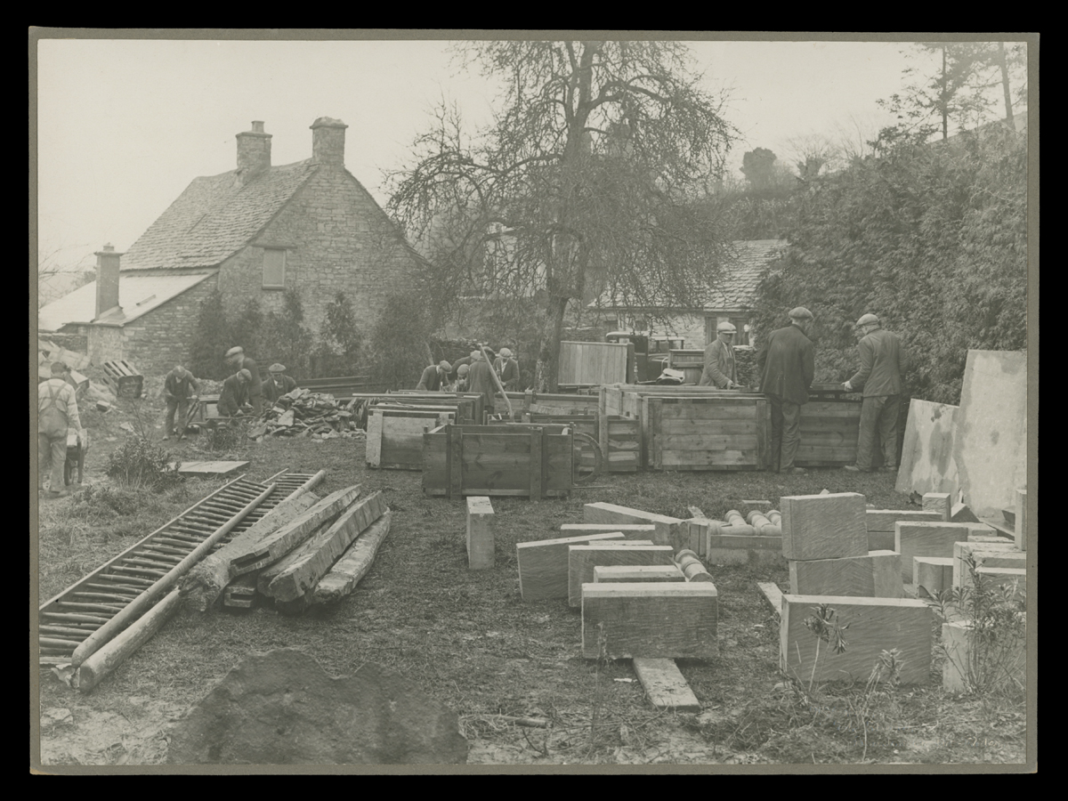 Dismantling Cotswold Cottage at its Original Site in England, 1929-1930 People around a group of crates and large stone building blocks in front of a stone house