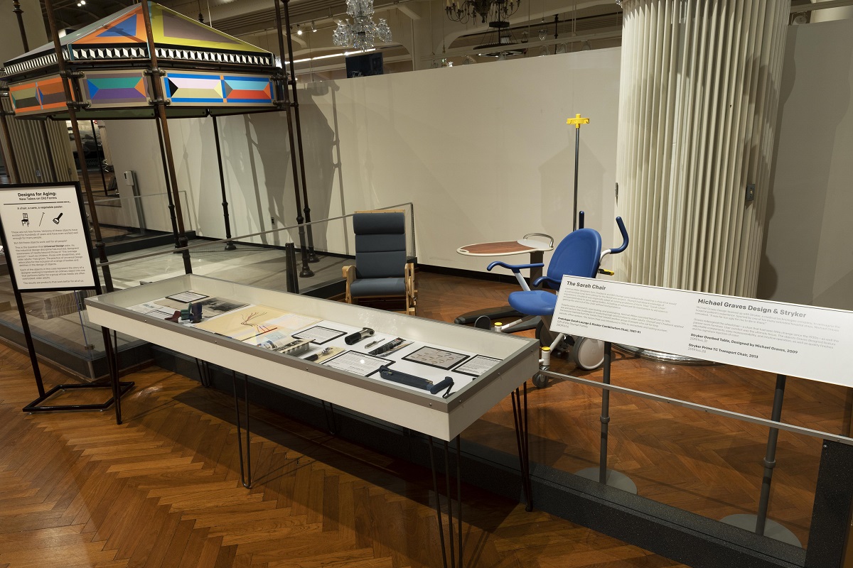 Designs for Aging: New Takes on Old Forms temporary pop-up exhibit during 2022 Museum display with large flat case containing paper and small items, a few large artifacts behind it, and extensive labels on either side
