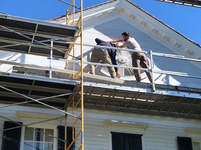 Noah Webster Home ellipse being reinstalled in the pediment Two men on scaffolding hold a large wooden architectural element on a building façade