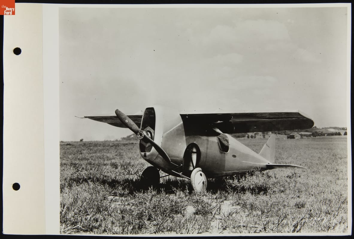 View of the Dayton Wright RB-1 Racer, August 1920 Black-and-white photo of small airplane parked in field with trees and buildings in the distance behind it
