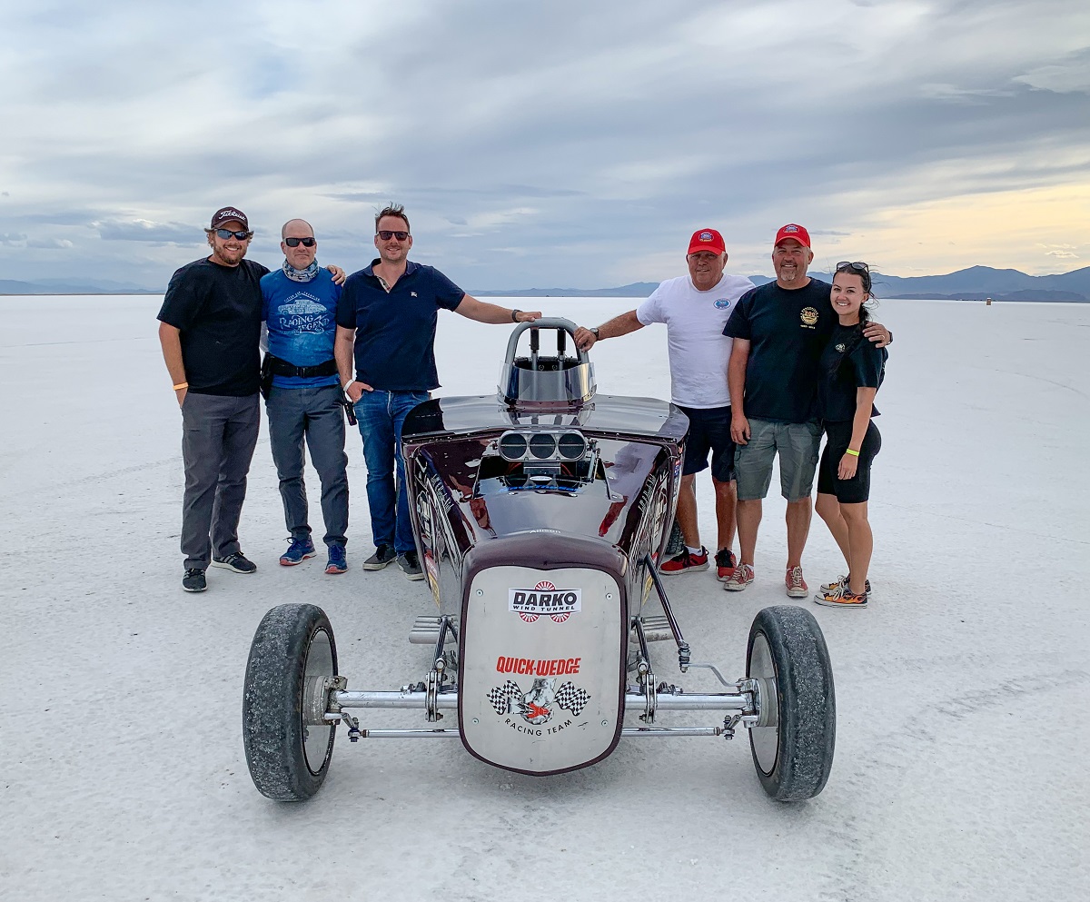 Some of the BRC Imagination Arts team with land speed racers Larry, Dallas, and Megan Volk. Six people stand around a race car on a salt plain with mountains in the background