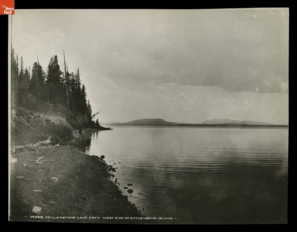 Yellowstone Lake from West Side of Stevenson's Island, Yellowstone National Park, 1870-1880 Black-and-white photo of a lake with a small shoreline with pine trees and a man fishing, with hills or mountains in the background