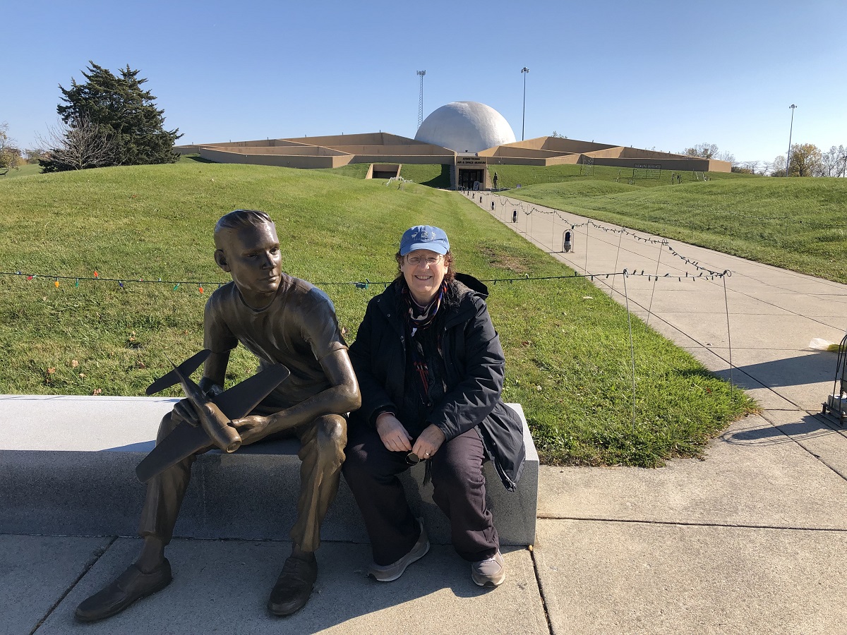 Donna R. Braden in front of the Armstrong Air & Space Museum Woman sits on bench next to statue of seated person, in front of a large lawn and low building