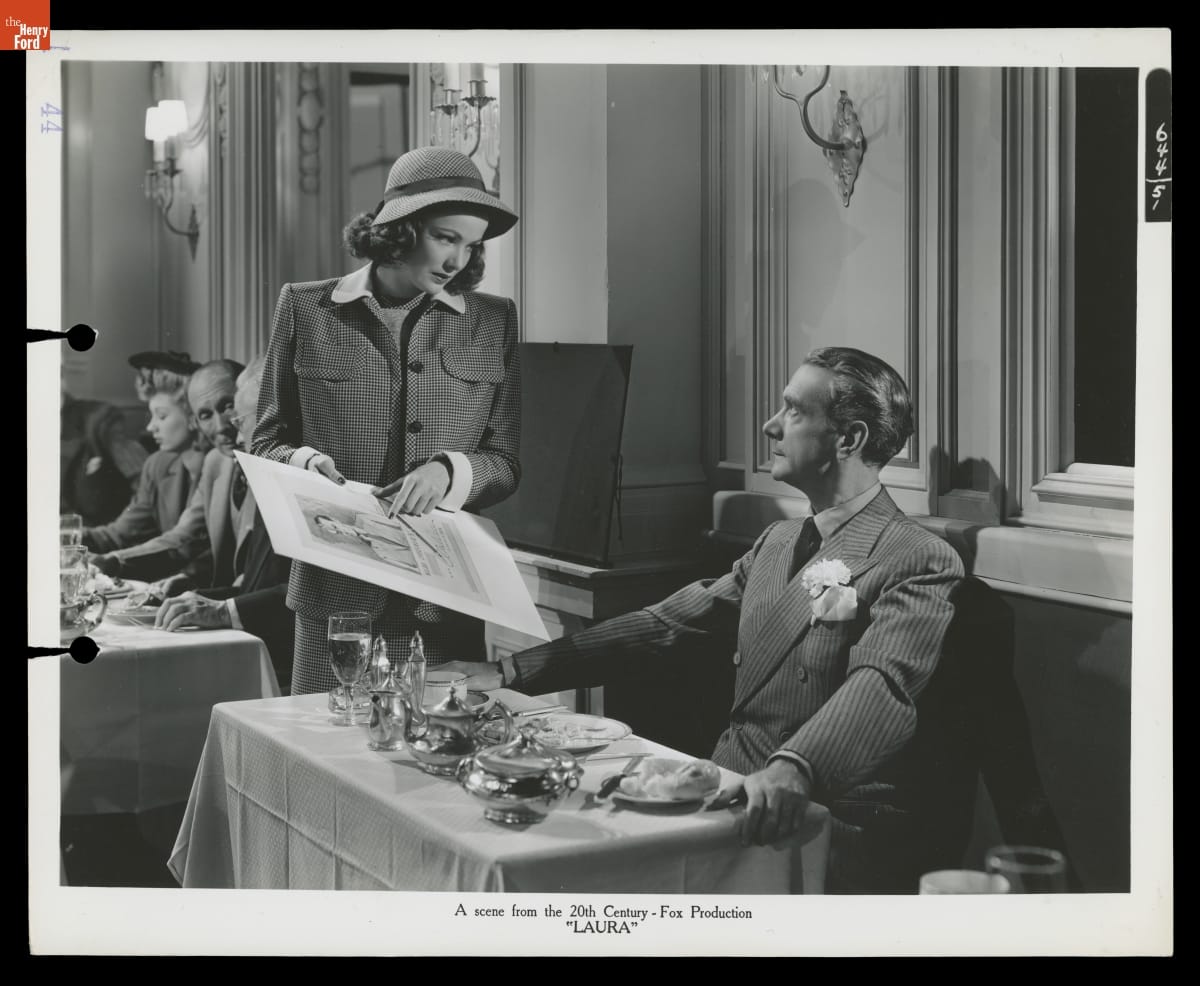 A Scene from the 20th Century-Fox Production "Laura" with Costume Designs by Bonnie Cashin, 1944 Black-and-white photo of a woman in a suit and hat standing at a table in a restaurant showing a poster to a man seated at a table