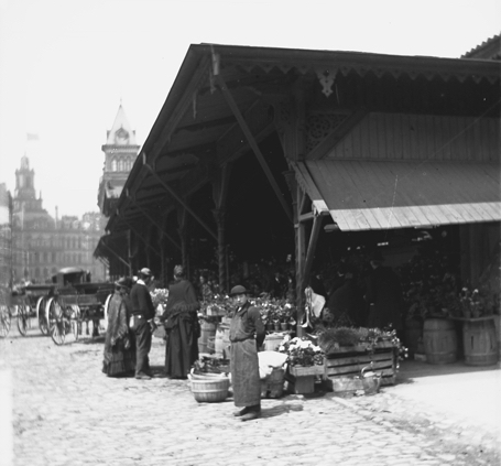 People look at flowers for sale at the Central Market in Detroit, Michigan People stand at edge of low open building, shopping at stands displaying flowers and other products