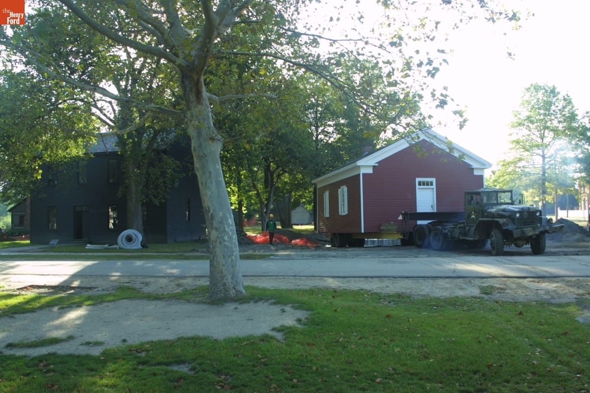 Dr. Howard's Office Being Relocated during the Greenfield Village Restoration Project, September 2002 Small red wooden building on trailer behind truck next to gray wooden building among lawns, roads, and trees