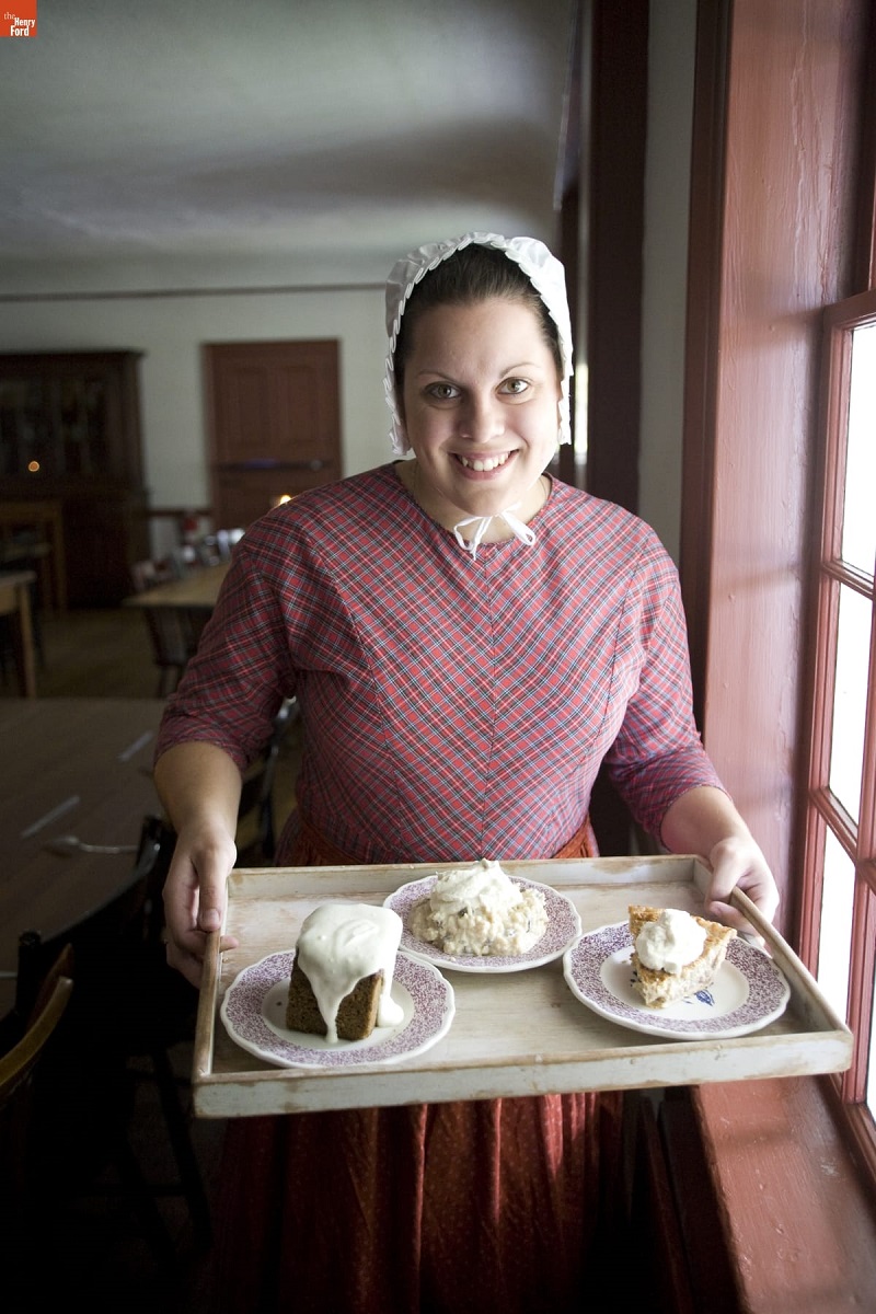 Eagle Tavern in Greenfield Village, October 2007 / Photographed by Michelle Andonian Woman in pink plaid dress and white bonnet smiles and holds tray of pastries