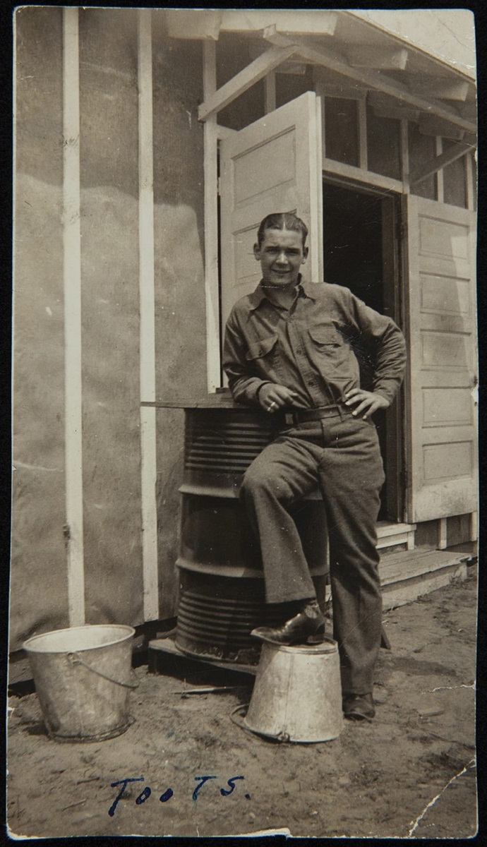 Stanley J. Zaleski at 1614th Co., Civilian Conservation Corps, Camp McComb, Munising, Michigan, April-September 1934 Man wearing uniform leans on piece of equipment with one foot on upturned bucket outside structure