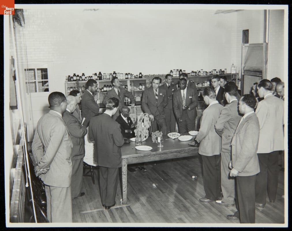 George Washington Carver at the Dedication of Carver Nutrition Laboratory, Dearborn, Michigan, 1942 Group of people stands around a large table in a room with shelves filled with many small bottles and jars