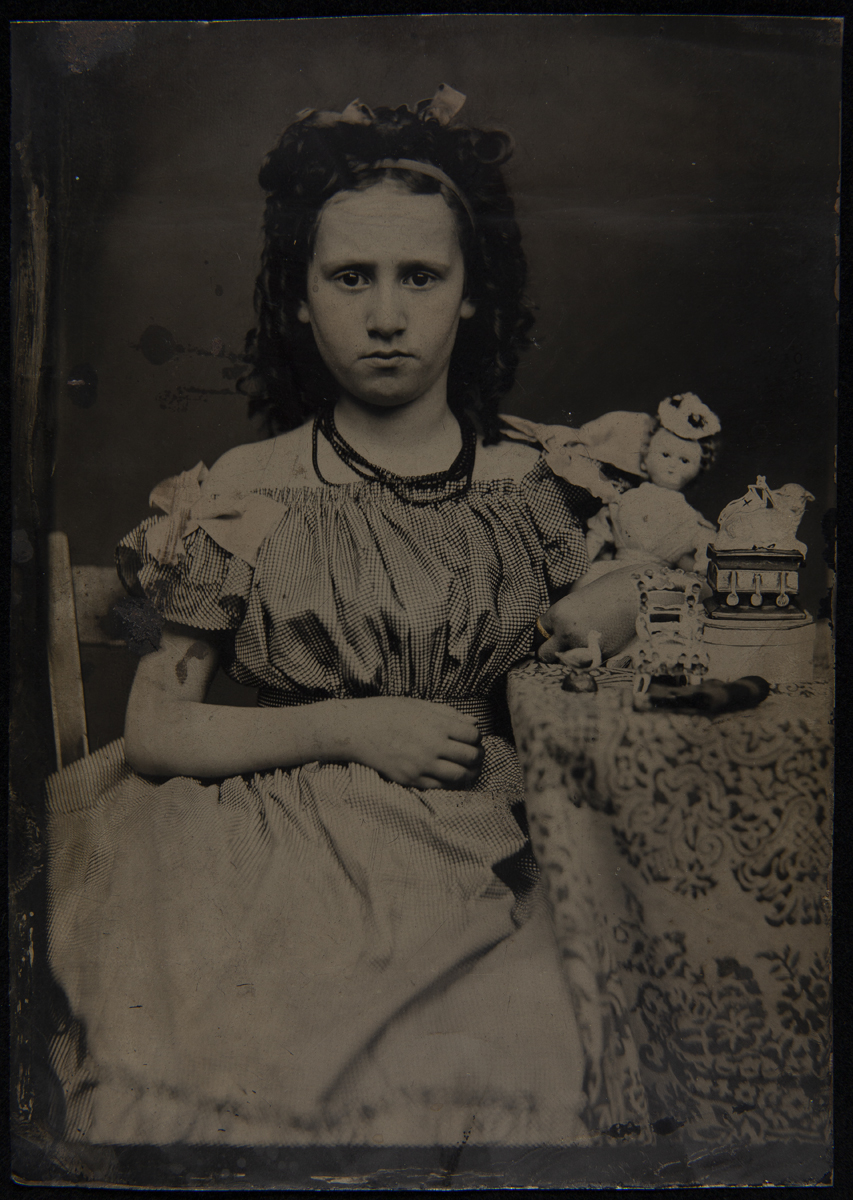 Girl Sitting at a Table with Her Toys, circa 1870 Girl in necklace and dress sitting by a table covered with a tablecloth with toys on top