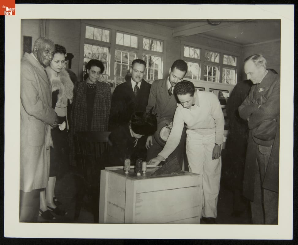George Washington Carver, Clara Ford, and Henry Ford at Dedication of George Washington Carver Museum, March 1941 Black-and-white photo of group of people standing around a box or crate filled with concrete