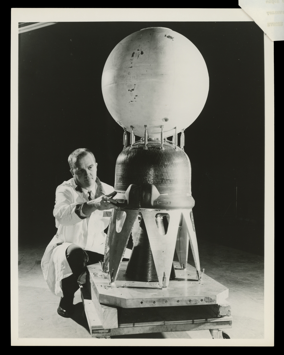 Lunar Capsule and Retrorocket for NASA Ranger 3 Spacecraft Being Assembled, April 1960 Man in lab coat kneels, working on a piece of equipment with stands, a dome-shaped middle portion, and a sphere at the top