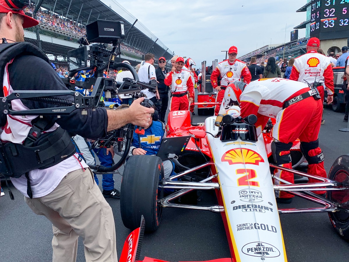 Filming Joseph Newgarden at the Indy 500. Man holding camera films long, low race car with pit crew in uniforms standing nearby and crowd in grandstands to both sides